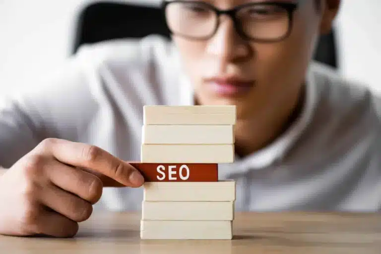 Person places a red 'SEO' block between stacks of small white books on a wooden desk, symbolizing SEO building blocks.